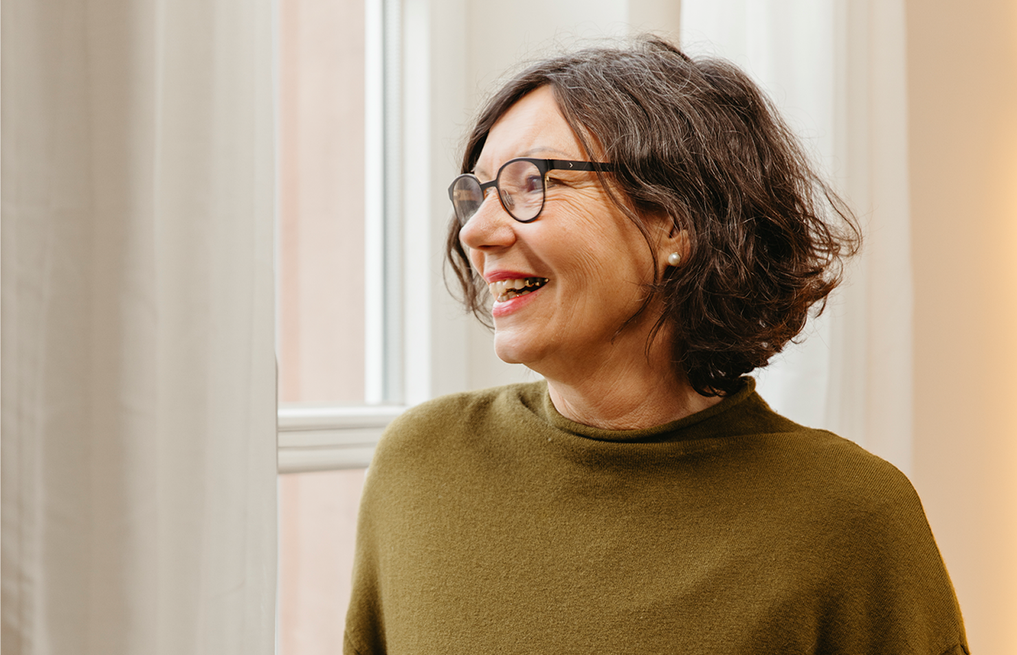 Eine Frau mit kurzen dunklen Haaren und Brille, die einen olivgrünen Pullover trägt, blickt lächelnd aus einem hellen Fenster mit weißen Vorhängen.
